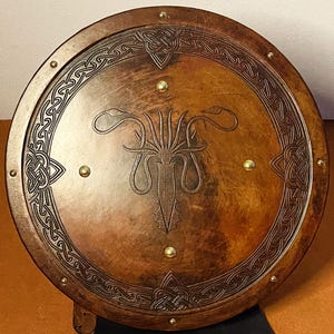 May include: A round, wooden shield with a brown finish and intricate Celtic knotwork around the edge. The shield features a central design and is accented with brass studs. The shield is a decorative item.