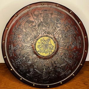 May include: A round, dark brown shield with intricate embossed designs of warriors on horseback and other figures. The shield has a gold-colored central emblem and is trimmed with brass studs, showcasing a historical or fantasy theme.