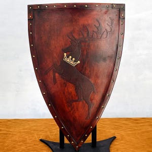Può includere: Scudo in legno marrone rossiccio scuro con un cervo incoronato d'oro in rilievo. Lo scudo è bordato da borchie in ottone e montato su un supporto nero. Oggetto decorativo.