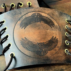 May include: Brown leather arm guard with an embossed owl design. The design features two owls with outstretched wings within a circle. The arm guard has eyelets with black cord lacing and gold-colored grommets.