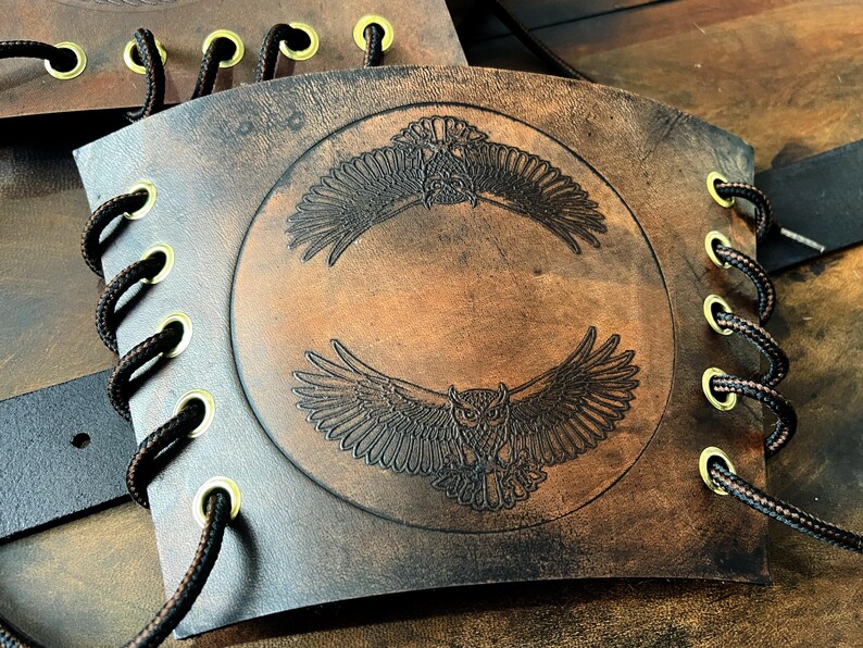 May include: Brown leather arm guard with an embossed owl design. The design features two owls with outstretched wings within a circle. The arm guard has eyelets with black cord lacing and gold-colored grommets.
