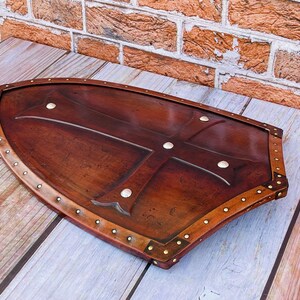 May include: A brown leather shield with a metal cross design and brass rivets around the edge. The shield is on a wooden surface.