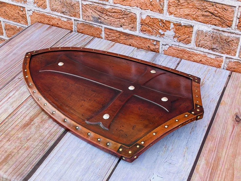 May include: A brown leather shield with a metal cross design and brass rivets around the edge. The shield is on a wooden surface.