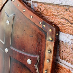May include: A brown leather shield with a cross design and brass rivets. The shield is decorated with a distressed finish and has a worn look.