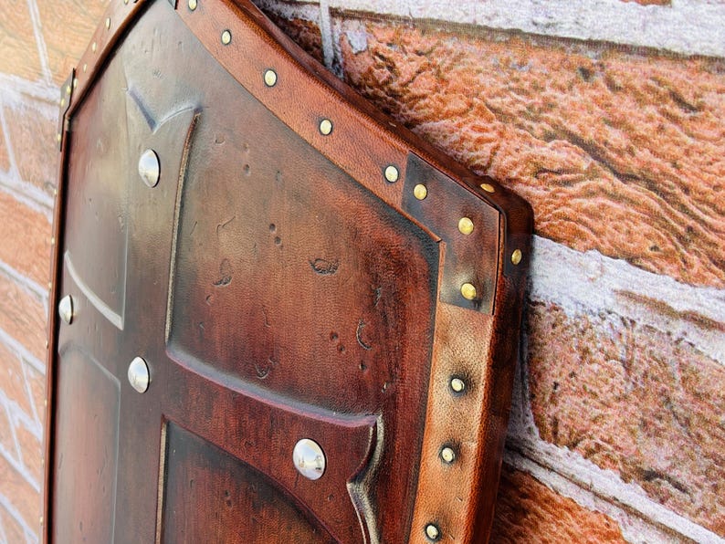 May include: A brown leather shield with a cross design and brass rivets. The shield is decorated with a distressed finish and has a worn look.