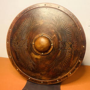 May include: A round, brown shield with an embossed design of two eagles facing each other. The shield has a central boss and is trimmed with small, evenly spaced rivets. The shield is made of wood.
