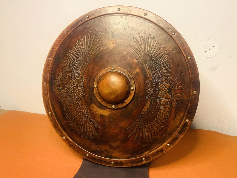 May include: A round, brown shield with an embossed design of two eagles facing each other. The shield has a central boss and is trimmed with small, evenly spaced rivets. The shield is made of wood.
