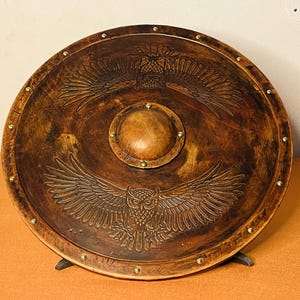 May include: A round, brown shield with an embossed owl design. The shield has a central circular boss and is adorned with brass-colored studs around the perimeter. The owl's wings are spread, and the shield is supported by a black stand.