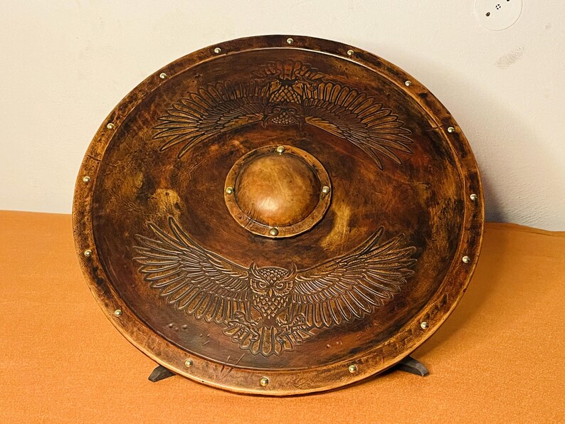 May include: A round, brown shield with an embossed owl design. The shield has a central circular boss and is adorned with brass-colored studs around the perimeter. The owl's wings are spread, and the shield is supported by a black stand.