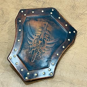 May include: A handcrafted, shield-shaped item with a distressed, aged appearance. The shield features a central emblem with a sword, wings, and geometric shapes, in shades of brown and blue. The edges are lined with small, gold-colored rivets.