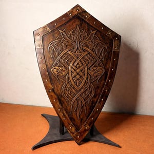 May include: A pointed wooden shield featuring a dark brown finish and intricate carved knotwork patterns. The shield is bordered by a wooden frame secured with evenly spaced metal rivets and is supported by a dark grey metal stand.