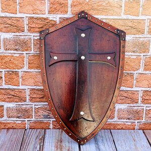 May include: A wooden shield with a cross design and metal accents. The shield is brown and has a distressed finish.