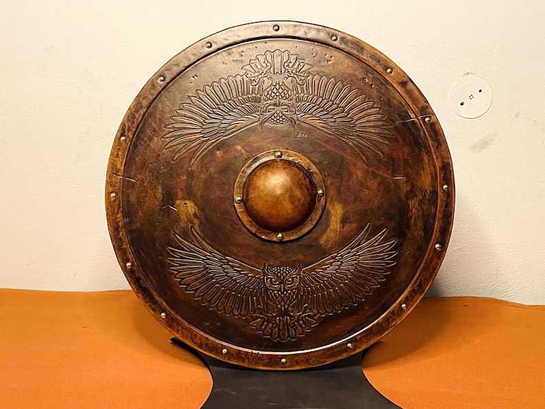 May include: A round, wooden shield with a detailed owl design. The shield is a dark brown color with a central boss and brass rivets around the edge. The owl design features outstretched wings.