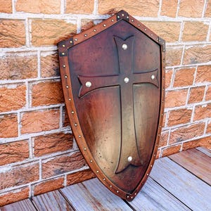 May include: A brown wooden shield with a metal cross design and silver studs around the edge. The shield is leaning against a brick wall.