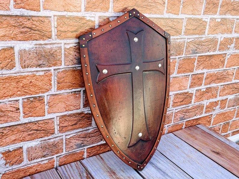 May include: A brown wooden shield with a metal cross design and silver studs around the edge. The shield is leaning against a brick wall.