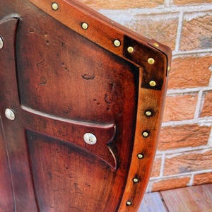 May include: A brown leather shield with a cross design and brass rivets. The shield is decorated with a distressed finish and has a wooden frame.