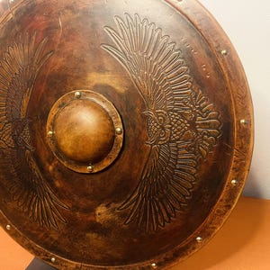 May include: A round, brown shield with a central, circular boss and embossed owl wing designs. The shield has a weathered appearance with brass-colored rivets around the edge. The shield is made of wood and is likely for decorative purposes.
