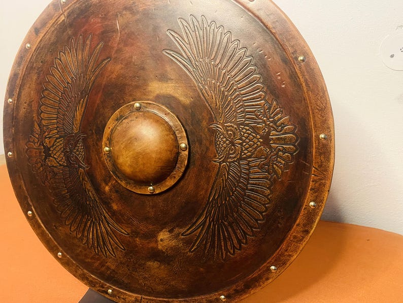 May include: A round, brown shield with a central, circular boss and embossed owl wing designs. The shield has a weathered appearance with brass-colored rivets around the edge. The shield is made of wood and is likely for decorative purposes.