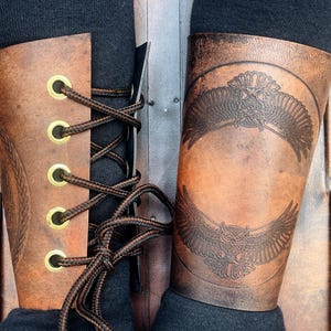 May include: Brown leather arm guards with an embossed eagle design. The arm guards have a laced closure with brass eyelets. The leather has a distressed finish, and the design is visible on the front.