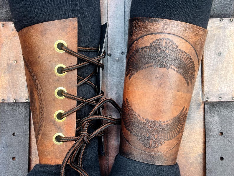 May include: Brown leather arm guards with an embossed eagle design. The arm guards have a laced closure with brass eyelets. The leather has a distressed finish, and the design is visible on the front.