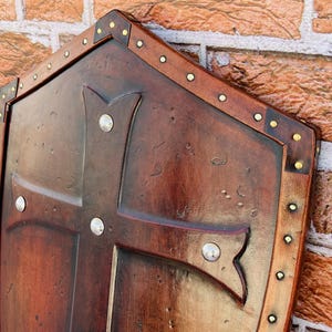 May include: A brown wooden shield with a cross design and metal accents. The shield is decorated with a distressed finish and has a leather trim.