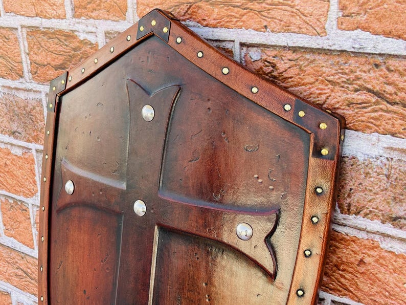 May include: A brown wooden shield with a cross design and metal accents. The shield is decorated with a distressed finish and has a leather trim.
