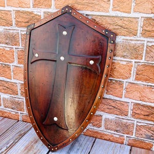 May include: A wooden shield with a cross design and metal accents. The shield is brown with a distressed finish and has a metal trim around the edge.