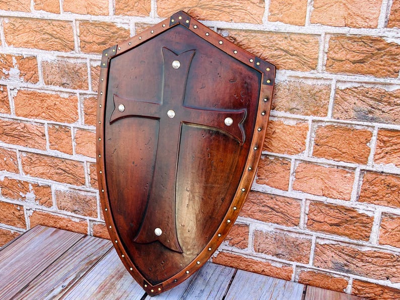 May include: A wooden shield with a cross design and metal accents. The shield is brown with a distressed finish and has a metal trim around the edge.