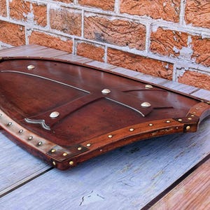 May include: A brown wooden shield with a leather trim and metal accents. The shield is decorated with a cross-shaped design.