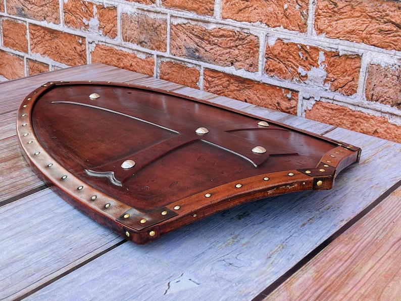 May include: A brown wooden shield with a leather trim and metal accents. The shield is decorated with a cross-shaped design.