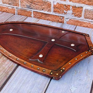 May include: A brown wooden shield with a cross design and brass accents. The shield is decorated with metal studs around the edge.