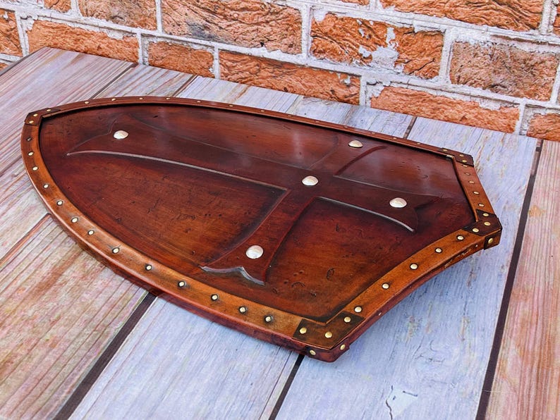 May include: A brown wooden shield with a cross design and brass accents. The shield is decorated with metal studs around the edge.
