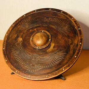 May include: A round, wooden shield with a brown finish, featuring an embossed owl and eagle design. The shield has a central boss and is accented with brass studs around the perimeter. It is supported by a black stand.