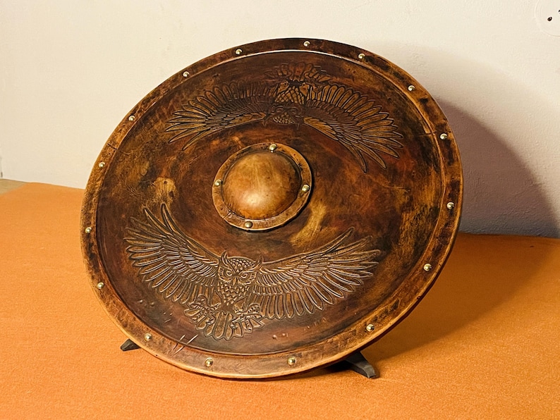 May include: A round, wooden shield with a brown finish, featuring an embossed owl and eagle design. The shield has a central boss and is accented with brass studs around the perimeter. It is supported by a black stand.