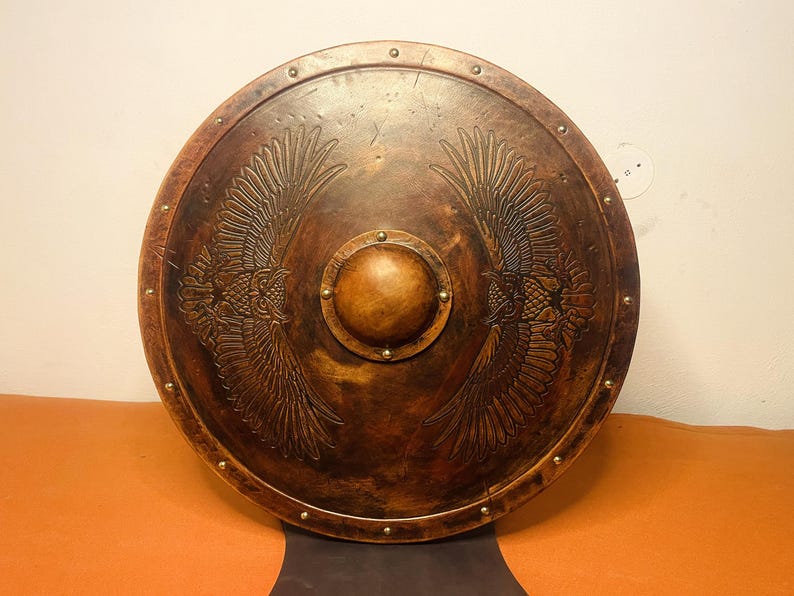 May include: A round, antique-style shield with a dark brown finish. The shield features an embossed design of two symmetrical eagle wings. A central, circular boss and brass rivets complete the design. This item is suitable for display or costume.