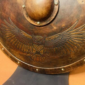 May include: A round, antique-style shield with a detailed owl design. The shield is primarily brown with a metallic finish and features a raised central boss and decorative rivets around the edge. The owl is depicted with outstretched wings.