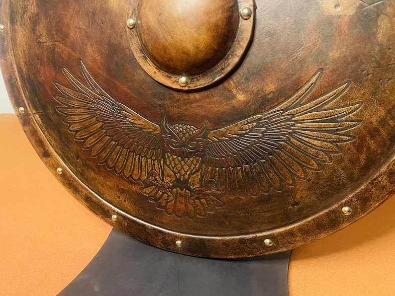 May include: A round, antique-style shield with a detailed owl design. The shield is primarily brown with a metallic finish and features a raised central boss and decorative rivets around the edge. The owl is depicted with outstretched wings.