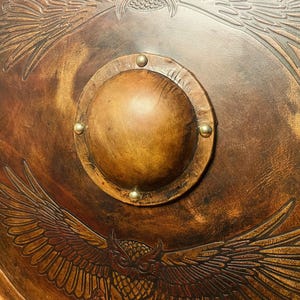May include: A round, brown shield with an embossed owl and eagle design. The shield has a central, raised boss secured with brass rivets. The overall color is a warm brown with darker shading, giving it an aged appearance.