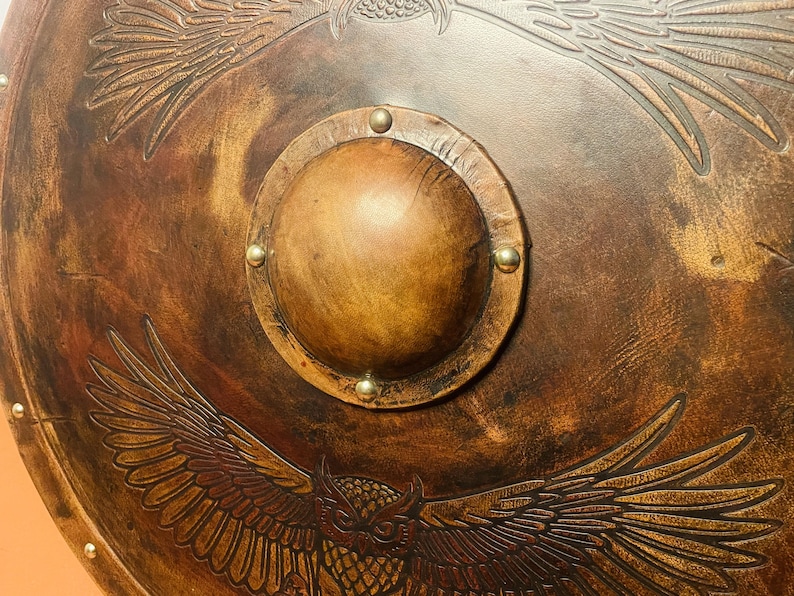 May include: A round, brown shield with an embossed owl and eagle design. The shield has a central, raised boss secured with brass rivets. The overall color is a warm brown with darker shading, giving it an aged appearance.