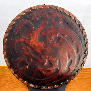 May include: A circular shield in dark brown, featuring a red dragon emblem. The shield has a textured, leather-like appearance, a decorative border, and brass studs. The dragon design is embossed on the shield's surface.