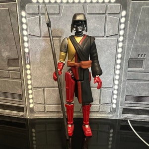 K&ouml;nnte beinhalten: Eine schwarze und rote Actionfigur mit einem schwarzen Helm und einem langen schwarzen Umhang. Die Figur h&auml;lt eine lange, d&uuml;nne, schwarze Lanze. Die Figur steht auf einem grauen Hintergrund.