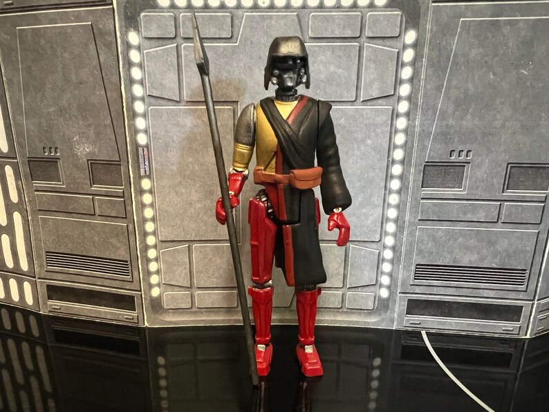 K&ouml;nnte beinhalten: Eine schwarze und rote Actionfigur mit einem schwarzen Helm und einem langen schwarzen Umhang. Die Figur h&auml;lt eine lange, d&uuml;nne, schwarze Lanze. Die Figur steht auf einem grauen Hintergrund.