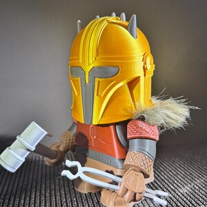 May include: A 3D-printed figure of a character in a yellow helmet with grey accents, wearing a red and brown outfit. The figure holds a white hammer and tongs. The figure has a fur collar.