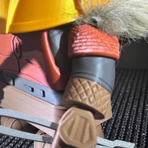 May include: Close-up of a detailed action figure. The figure has a yellow helmet with faux fur trim, red armour, and brown gauntlets. The figure is holding two silver-coloured weapons.