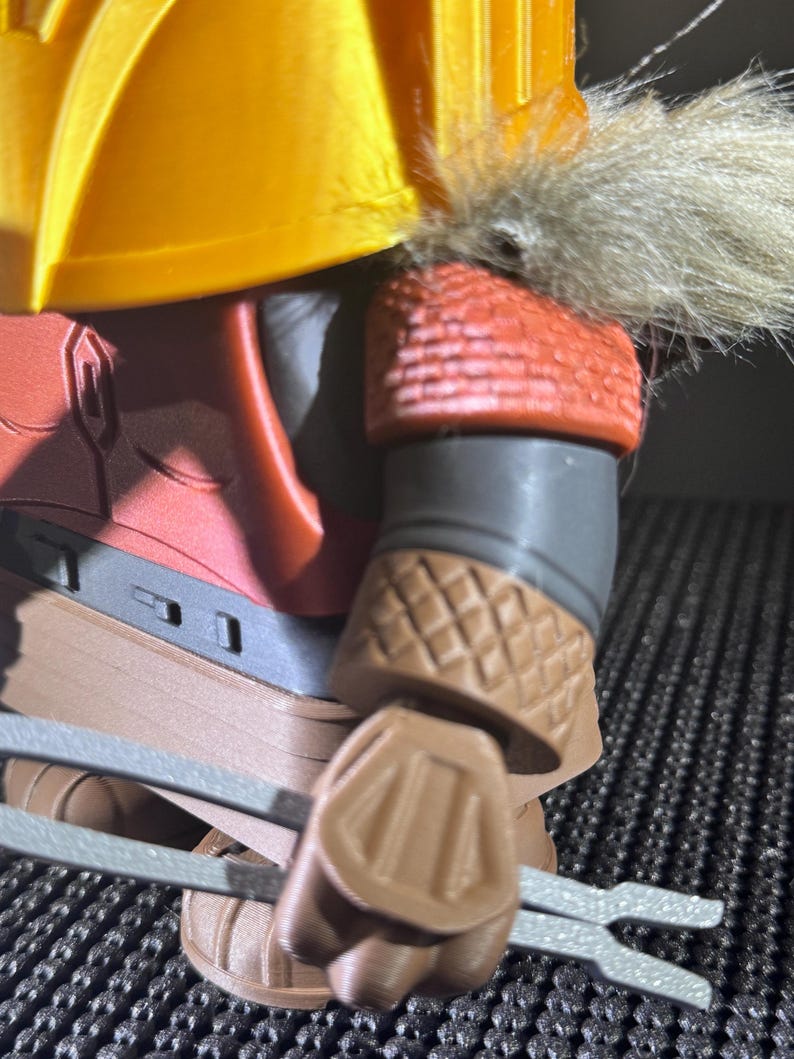 May include: Close-up of a detailed action figure. The figure has a yellow helmet with faux fur trim, red armour, and brown gauntlets. The figure is holding two silver-coloured weapons.