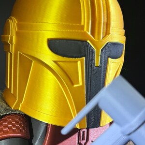 May include: A close-up of a gold Mandalorian helmet with black detailing. The helmet features a T-shaped visor and a textured finish. The image also shows a portion of the red and grey armour.