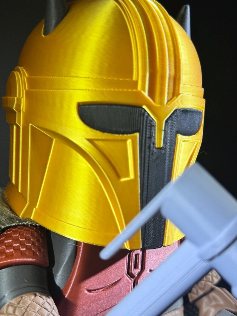 May include: A close-up of a gold Mandalorian helmet with black detailing. The helmet features a T-shaped visor and a textured finish. The image also shows a portion of the red and grey armour.