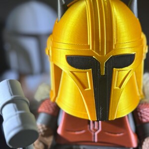 May include: A gold-coloured helmet with black detailing, reminiscent of a science fiction character. The helmet is part of a figurine, with a red and brown torso and grey arms. A grey hammer is held in the figurine's hand.