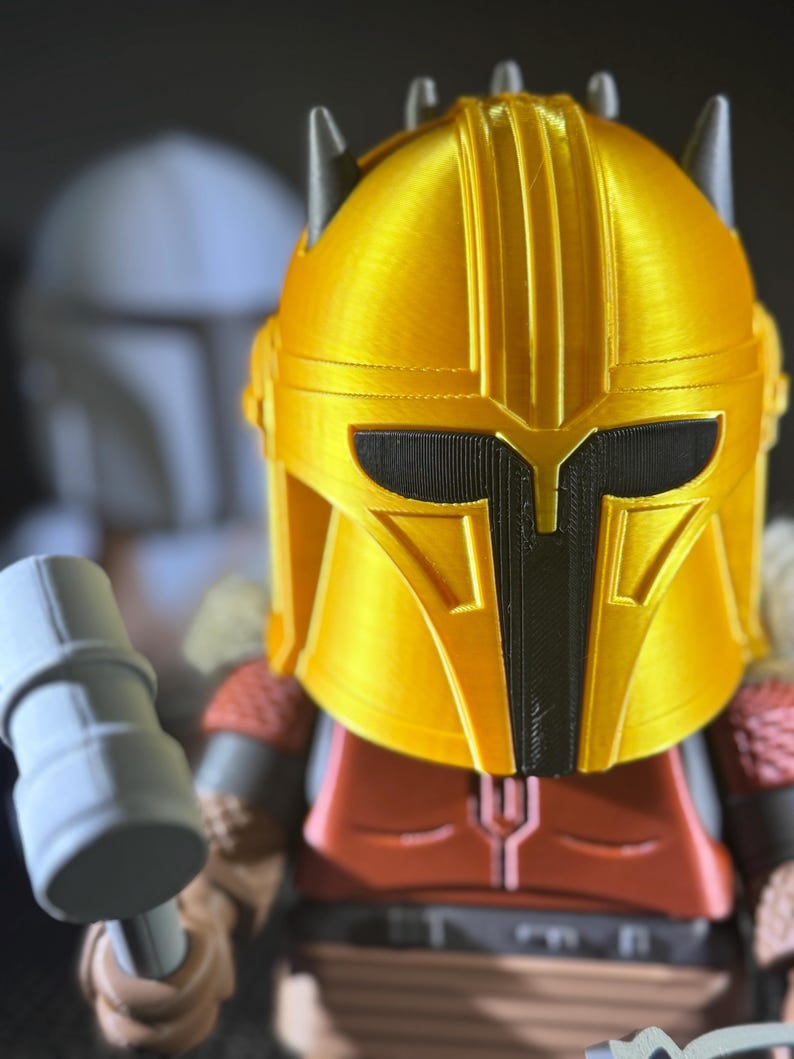 May include: A gold-coloured helmet with black detailing, reminiscent of a science fiction character. The helmet is part of a figurine, with a red and brown torso and grey arms. A grey hammer is held in the figurine's hand.