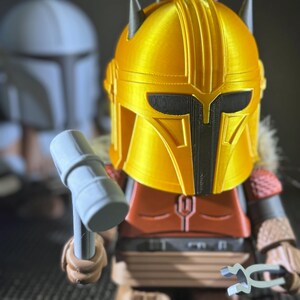 May include: A 3D-printed figure of a character from a science fiction series. The figure has a gold helmet, red and brown armour, and holds a hammer and a tool. Another figure is in the background.